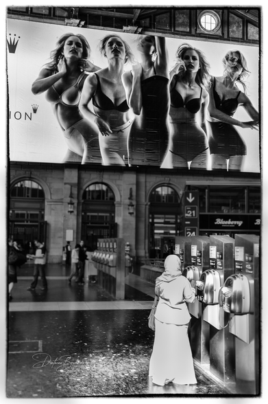 Das Schwarz-Weiss-Bild zeigt eine Szene in einem Bahnhof. Im oberen Bereich des Fotos dominiert ein riesiges Werbeplakat mit mehreren Frauen in Unterwäsche, die dramatisch posieren. Ihre Gesichter und Körper sind ausdrucksstark beleuchtet, was ihnen eine fast plastische Wirkung verleiht. Im unteren Bereich steht eine Frau mit Kopftuch und langem Mantel an einem Münztelefon, während andere Personen unscharf durch die Bahnhofshalle laufen. Die Kombination aus der dramatischen Werbung und der alltäglichen Szene schafft einen starken Kontrast und lädt zum Nachdenken ein.