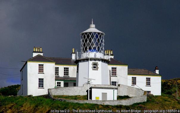 Lit by low sunlight under a sky black with storm lids a vivid white lighthouse and associated buildings