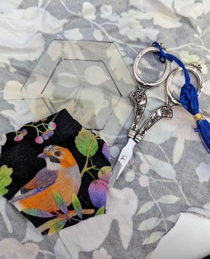 Stoffrückseite, ein florales Motiv mit verschiedenen Tieren (Hasen, Bären, Vögel) schimmert durch. Mit Bleistift sind Hexagone auf den Stoff gezeichnet. Eines ist schon ausgeschnitten und liegt mit der richtigen Seite nach oben da, ein Rotkehlen ist hierauf zu sehen. Daneben eine verzierte, silberne Stickschere und ein Plastikhexagon aus dicker, durchsichtiger Folie, mit dem eins Fuzzy Cut machen kann.