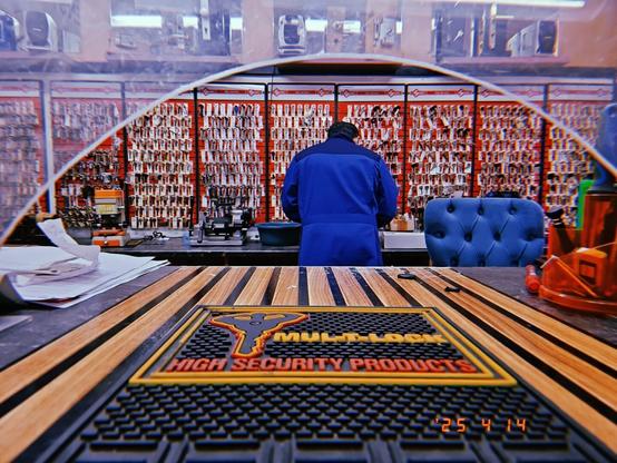 A locksmith in a blue coat working in front of a wall of keys pictured through the counter window.