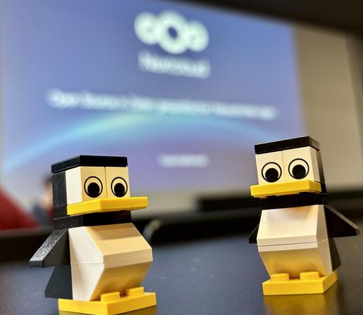Zwei kleine Pinguinfiguren aus LEGO stehen auf einem schwarzen Tisch. Sie sind stilisiert, mit großen, runden Augen und gelben Schnäbeln und Füßen. Im Hintergrund ist eine unscharfe Projektion zu sehen, auf der das Logo von Nextcloud sowie ein verschwommener Schriftzug mit dem Thema „Open Source“ erscheint. Die Atmosphäre wirkt wie in einem Konferenz- oder Workshopraum. Die beiden LEGO-Pinguine stehen im Vordergrund scharf und im Fokus.