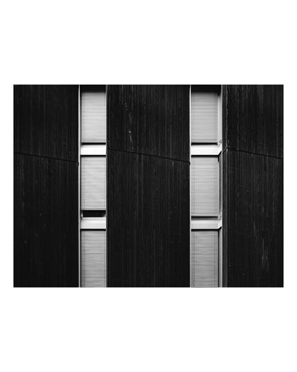 Black and white photo of a modern building facade featuring tall, narrow windows with closed blinds, set against dark vertical wooden panels. The strong lines and repetitive pattern create a minimalist, geometric architectural look.