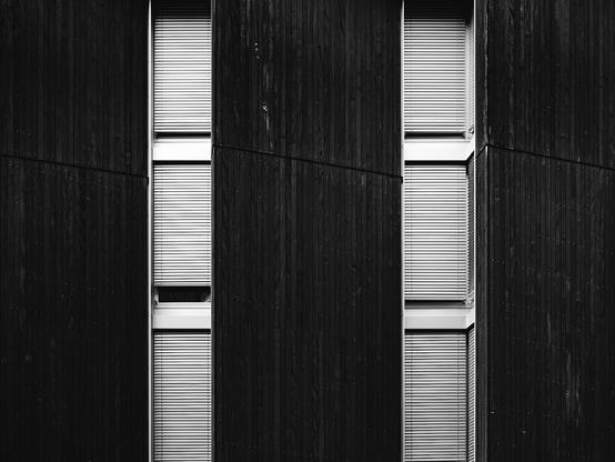 Black and white photo of a modern building facade featuring tall, narrow windows with closed blinds, set against dark vertical wooden panels. The strong lines and repetitive pattern create a minimalist, geometric architectural look.