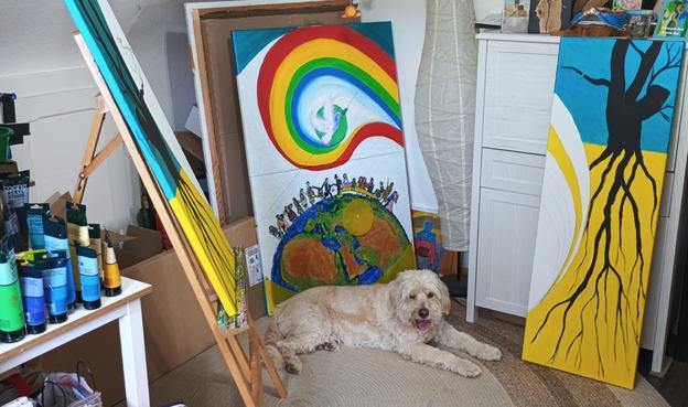In einem Atelier stehen drei nur teilweise erkennbare und noch unvollendete Gemälde auf einer Staffelei und auf dem Fußboden. Davor liegt ein freundlicher Hund mit hellem, langen Fell und total süßen Augen.