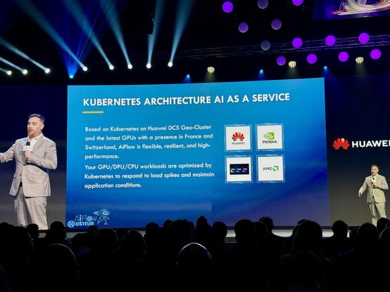 A man in a suit is speaking on a stage, holding a microphone. On the large screen behind him, it says "KUBERNETES ARCHITECTURE AI AS A SERVICE." Details follow about the use of Kubernetes on Huawei DCS Geo-Cluster and GPU technologies, with logos of Huawei, NVIDIA, Apple, and AMD. On the right side of the screen, the Huawei logo is visible. The audience is seated in the dark in front of the stage.