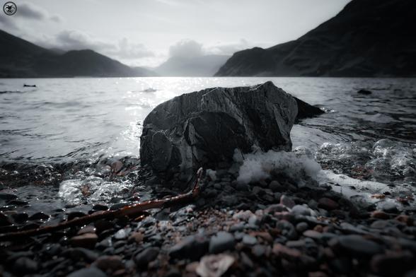monochrome shot of lake edge with rock in centre of shot, mountains in background