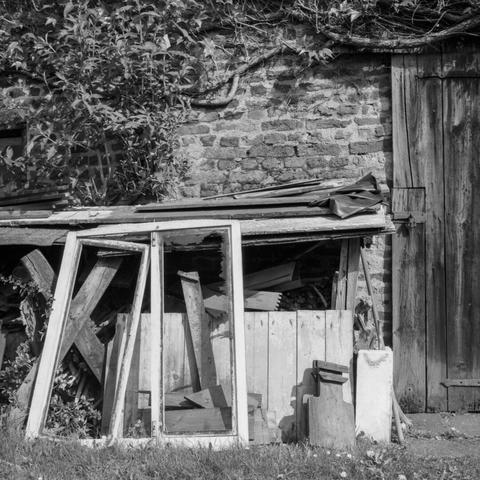 Black and white photo of a stack of assorted junk, like old planks of wood and winow frames next to a brick wall that is partially overgrown with ivy.