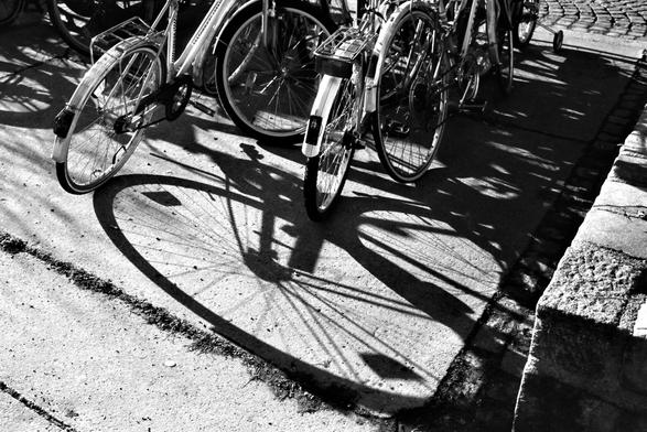 The black and white image shows a group of bicycles parked together, casting long, intricate shadows on the pavement, with two bike’s circular wheel shadows prominently forming a heart-like shape.