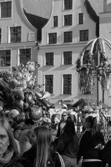 The image captures a street scene in Rostock, Germany. A crowd is gathered, seemingly enjoying a street fair or celebration. A large assortment of character balloons is prominently displayed in the foreground, while an ornate archway adorned with flowers and decorations stands nearby. The backdrop features a building and a Vodafone sign, adding to the urban atmosphere.