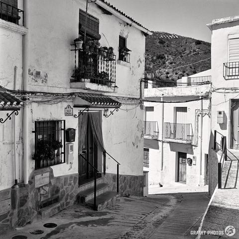 A black and white photo captures a narrow, hilly street in Torvizcón, Spain, lined with whitewashed buildings. Balconies adorned with plants overlook the alley below, while distant hills paint a picturesque backdrop.