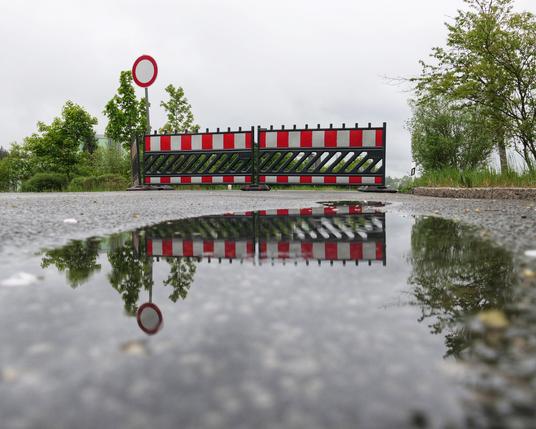 Das Bild zeigt eine Straßensperre mit rot-weißen Absperrbalken, die eine Straße blockieren. Im Vordergrund befindet sich eine Pfütze, die die Absperrbalken und die umliegenden Bäume reflektiert. Ein rundes Verkehrsschild mit einem roten Rand und weißem Innenbereich ist ebenfalls sichtbar, was auf ein Verbot der Durchfahrt hinweist. Die Umgebung ist grün mit Bäumen und Sträuchern, und der Himmel ist bewölkt. Die Perspektive des Bildes ist niedrig, fast auf Straßenniveau, was die Reflexion in der Pfütze besonders hervorhebt.