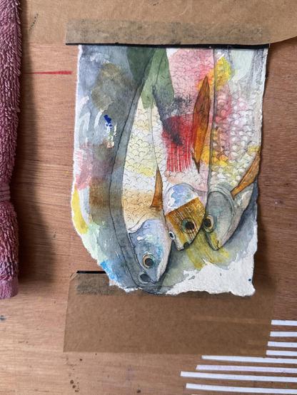Ein kleines Aquarellbild, das 4 Fische darstellt, umgeben von abstrakten Farben und Formen. Das Kunstwerk ist auf einer Holzoberfläche angebracht.