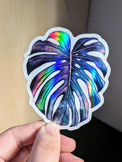 Photo of a left hand holding a diecut sticker. The sticker is a holographic monstera leaf drawing.