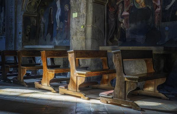 Sunlight spills in through a window and onto some of the wooden pews of the church of Santa Maria Nuova, a Romanesque church in Viterbo Italy. Murals depicting religious figures adorn the stone walls, adding a sense of reverence and antiquity to the scene.