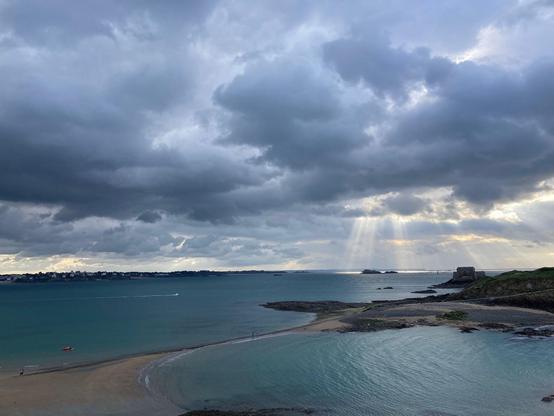 Vue côtière d'une plage bretonne sous un ciel nuageux dramatique. Des rayons de soleil percent les nuages, éclairant l'horizon marin. À droite, un fort historique se dresse sur un îlot rocheux, entouré par des eaux turquoise et calmes. Quelques promeneurs marchent sur le sable, ajoutant une touche de vie à ce paysage paisible et majestueux