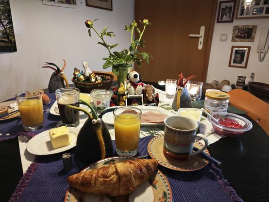 Frühstückstisch mit Päonien, Auswahl an Wurst, Käse, Krabben, Honig, Marmelade, gekochte Eier (unter Filzhühnern), Orangensaft, Kaffee und Croissants. Auf dem Tisch sitzen Snoopy, Woodstock, ein kleines Plüschmammut.