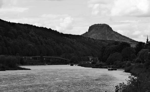 Schwarz-weiß Landschaftsfotografie. Vom Vordergrund fließt ein Fluss, der in der Ferne zwischen den Talflanken verschwindet. Im unruhigen Wasser spiegelt sich der teilweise bedeckte Himmel weiß. Die Ufer sind bedeckt von Büschen und Bäumen, zwischen denen vereinzelt Gebäude oder Infrastruktur wie Straßenlaternen zu sehen sind. In unterschiedlicher Entfernung liegen Boote an Anlegern, gegenüber vom Betrachter sieht man eine Brücke. Im Hintergrund steht ein Tafelberg eckig aus der Landschaft hervor. Durch die diesige Luft und schwache Sonnenstrahlen ist er heller als die nahen Talwände, jedoch steht auch er dunkel vor dem hellgrauen Wolkenhimmel hervor. Am rechten Bildrand ragt die Spitze eines Kirchturmes aus dem Wuchs hervor.