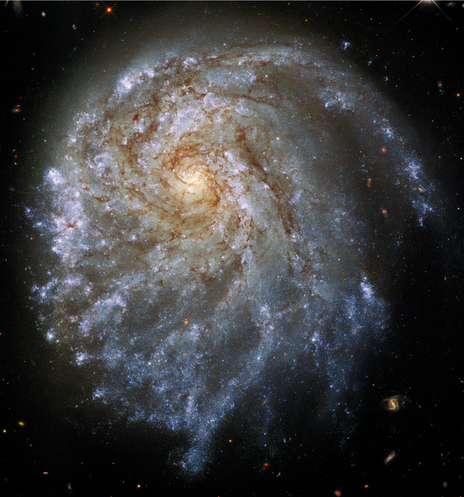Against the black background of space, which has distant stars and galaxies scattered across, is a large spiral galaxy filled with streaks of blue and pink. The galaxy has warped spiral arms that wrap clockwise around its bright yellow core. Toward the upper right, one of its arms appears to be breaking away from the rest of the galaxy.