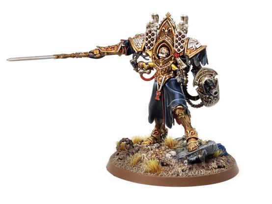 Photo of a Games Workshop Morvenn Vahl miniature - a figure in giant, baroque, golden armour wielding a spear and gun/shield combo - standing amongst a ruined battlefield.
