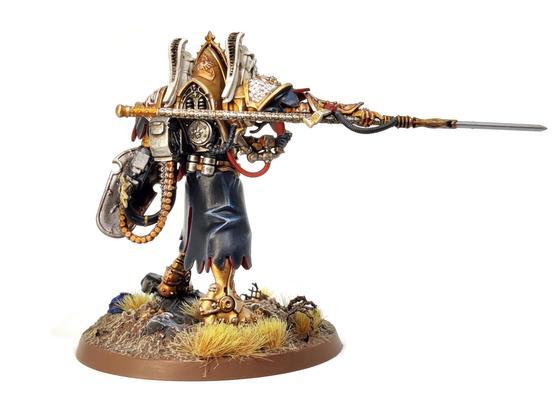 Photo of a Games Workshop Morvenn Vahl miniature - a figure in giant, baroque, golden armour wielding a spear and gun/shield combo - standing amongst a ruined battlefield.