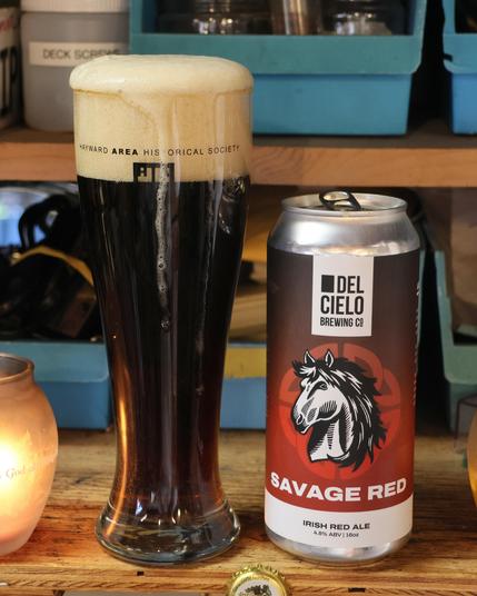 A pilsner glass filled with a dark amber fluid a topped with cream-colored foam stands next to a beer can. The can is themed in red and block, with a sketch of a proud-looking horse's head. The label reads “Del Cielo Brewing Company Savage Red, Irish Red Ale.”