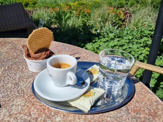 Eine Tasse Espresso und ein Glas Wasser auf einem kleinen Tablett auf einem Tisch in der Sonne, daneben ein Pappbecher mit einer Kugel Schokoladeneis. Im Hintergrund ist eine gepflegte Rabatte zu erkennen.