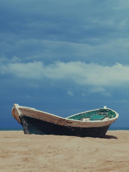 A weathered wooden boat with peeling green and white paint sits abandoned on a sandy beach under a dramatic blue sky filled with layered clouds. The sea is barely visible on the horizon, creating a sense of solitude and timelessness.