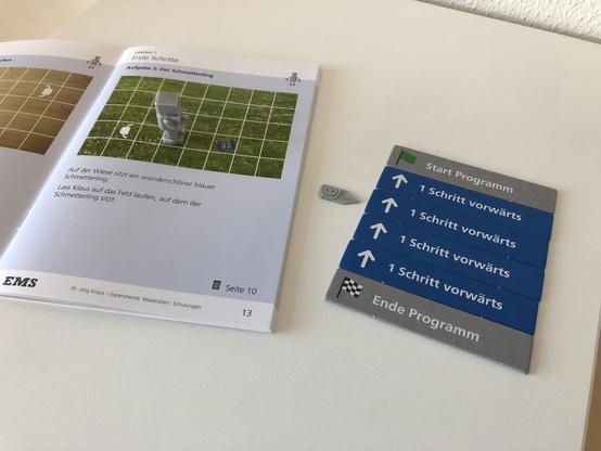 Das Bild zeigt einen kleinen grauen 3D-gedruckten Roboter, der auf einem Raster/Plan in einem gedruckten Heft steht. Daneben liegen Karten mit Programmierblöcken (Starte Programm, 1 Schritt vorwärts usw.).