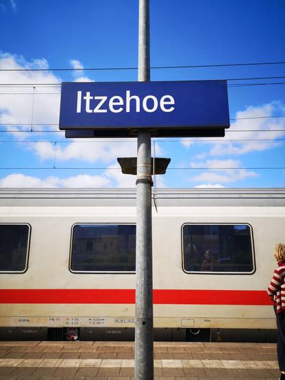 Abschnitt eines ICs unter blauem Himmel mit ein paar weißen Wolken, davor das Schild Itzehoe auf dem Bahnsteig, eine Person rechts kommt gerade ins Bild.