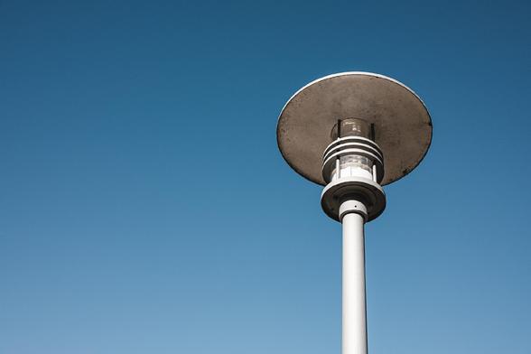 Minimalist photo of a tall, modern street lamp with a wide circular top against a clear blue sky. The lamp's industrial design contrasts with the smooth, cloudless backdrop.