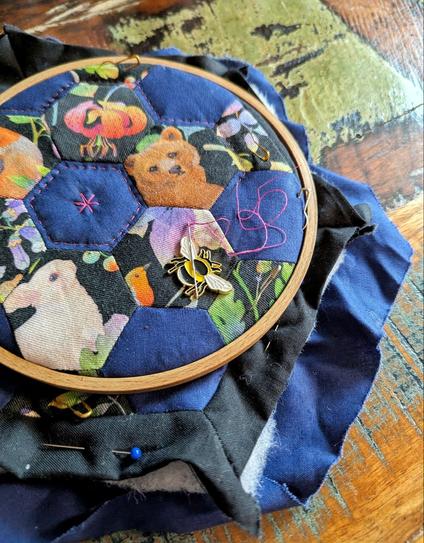 Dark table. On the table lies a embroidery hoop, filled with a small quilting project in black, dark blue and some hexies with woodland critters and plants. In the middle you can see a classic hexagon epp flower. On the outside you can see the different layers of the small quilt sandwich with top, binding and back. It's quilting in Progress with pink quilt thread. The needle lies on a needle magnetic minder in form of a honey bee.