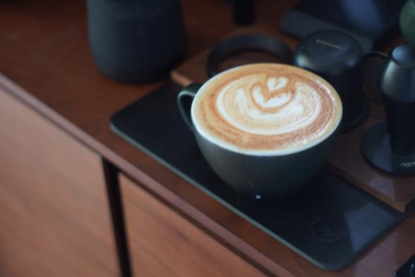 Slightly blurry textured black porcelain cup of coffee with latte art. The surface of the coffee is speckled with cinnamon. It sits on a black leather tamp mat on a warm walnut sideboard surrounded by matte black coffee gear.