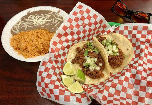 It's two tacos with lime on a white and red piece of paper with some rice and beans behind the tacos