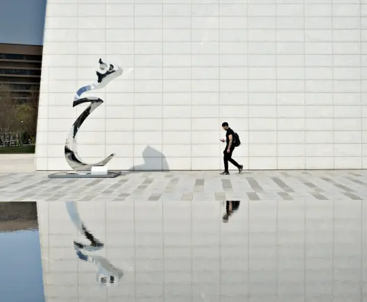 A minimalist architectural photograph showing the exterior of the Aga Khan Museum in Toronto. A large, pristine white wall with square marble tiles forms the backdrop. In the foreground, a shiny abstract metal sculpture with curved, flowing lines stands on the left side of the frame. A person wearing black clothing and carrying a backpack walks past the sculpture while looking at their phone. The scene is reflected in a shallow reflecting pool at the bottom of the image, creating a mirror effect of both the sculpture and the pedestrian. The composition emphasizes clean lines, geometric patterns, and the interplay between the modern architecture, contemporary art, and human presence.