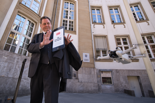 Ein Mann mit Koffer (es ist padeluun) hält ein überdimensioniertes Smartphone, auf dem steht "DB Schnüffelnavigator". Neben ihm fliegt eine Drohne. Er hält die Hände abwehrend hoch. Im Hintergrund ein Gerichtsgebäude.