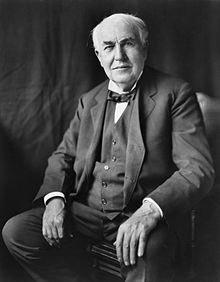 Thomas Edison sitzt auf einem Stuhl. Er trägt einen dunklen Anzug und eine Fliege. Seine Hände ruhen entspannt auf seinen Oberschenkeln, und er schaut direkt in die Kamera. Der Hintergrund ist unscharf und eher neutral.
