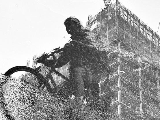 A black and white image depicting a reflection of a person riding a bicycle, distorted in water, with a construction site visible in the background.