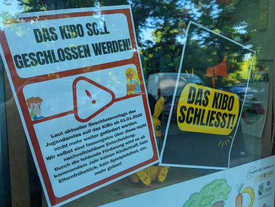 Zwei Plakate im Schaufenster. Die Scheibe spiegelt, Plakat links: "Das Kibo soll geschlossen werden" und kleinerer Text. Das Plakat rechts: "DAS KIBO SCHLIESST!" - ganz groß.