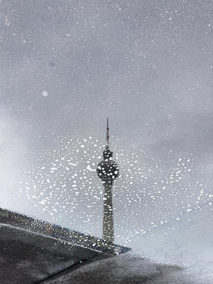 An abstract view of a tall tower, partially obscured by bubbles and streaks of water, set against a gray, overcast sky. The reflection appears on a wet surface, creating a unique visual effect.