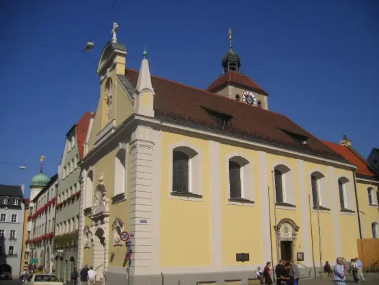 The image depicts a building with a distinctive architectural style, featuring a light-colored façade and a prominent clock tower. It seems to be a historical or significant building, likely located in a European city, given the design elements. The surrounding area includes other buildings and a clear blue sky. There are also people visible, indicating that it might be a public space.

Image Credits: Wikimedia / Szeder László / CC BY-SA 4.0