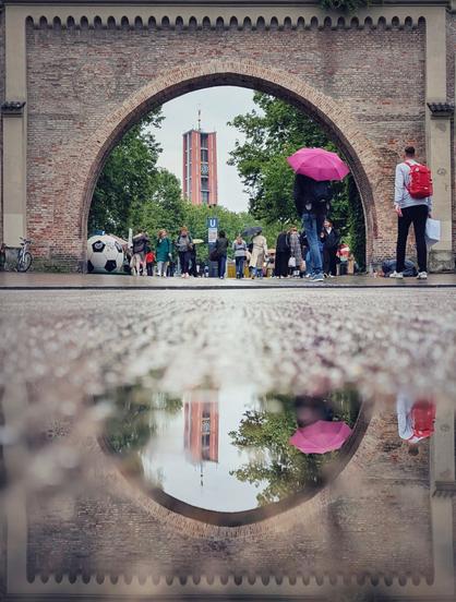 Das Bild zeigt eine beeindruckende städtische Szene, die durch einen großen, alten Backstein-Torbogen eingerahmt wird. Durch diesen Bogen hindurch erkennt man eine Gruppe von Menschen, einige mit Regenschirmen, die über einen Platz schlendern. In der Ferne erhebt sich ein hoher Turm mit einer Uhr. Im Vordergrund spiegelt sich der Turm sowie der Bogen in einer Pfütze. Links neben dem Bogen steht ein großes Fußballmodell. Vor dem Tor befinden sich rechts eine Person mit einem pinken Regenschirm und ein Mann mit einem roten Rucksack.