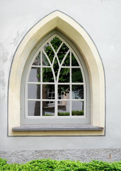 Ein kleines gotisches Fenster, das durch zwei unterteilte, verbundenen Spitzbogenrahmen aus weißem Holz in 20 Einzelscheiben unterteilt wird und zusätzlich außen grau umrahmt ist. Darunter sieht man eine steinerne Fensterbank. Die hellgrau gemalte Fensterlaibung hat eine abgeschrägte Kante, die umgebende Mauer ist hellgrau, am unteren Rand rauh. Darunter sieht man einen schmalen Streifen einer grünen Hecke.

A small Gothic window, which is divided into 20 individual panes by two divided, connected pointed arch frames made of white wood and is also framed in gray on the outside. A stone window sill can be seen underneath. The light gray painted window reveal has a beveled edge, the surrounding wall is light gray, rough at the lower edge. A narrow strip of green hedge can be seen below it.