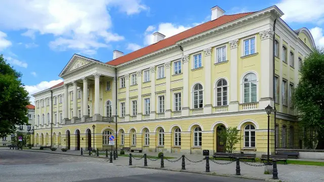 The image depicts a large, neoclassical building characterized by its yellow façade, white accents, and a red-tiled roof. It features several large windows and pillars at the entrance. The surrounding area includes trees, benches, and a clear blue sky. This style of architecture is often seen in historical or governmental buildings.

Image Credits: Wikimedia / Bartosz MORĄG / CC BY-SA 3.0