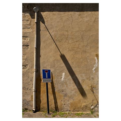 Flat image of a building with a cracked, weathered stone wall. On the left, a pipe runs from the bottom all the way to the top of the wall. A pole with a dead end sign is right next to it, leaning towards the pipe.  A streetlamp which is not visible casts a long, diagonal shadow from the top left to the bottom right.