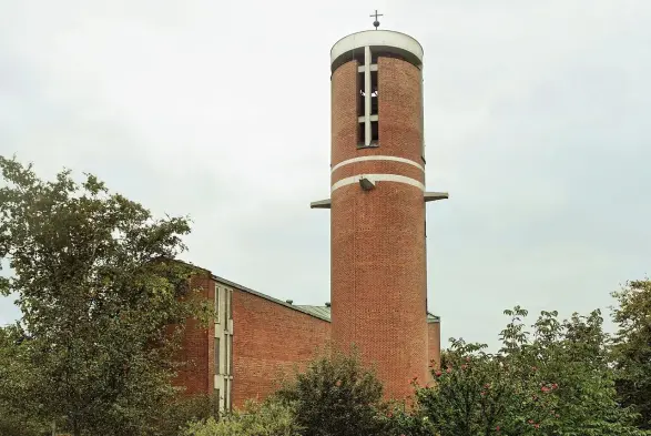 The image appears to depict a brick building with a tall, round tower. The tower features a cross at the top and has several openings, possibly for bells. The building itself seems to have a modern architectural design and is surrounded by greenery, including trees and plants. The sky is overcast, giving the scene a muted appearance.

Image Credits: Wikimedia / Mailtosapderivative work Hic et nunc / CC BY-SA 3.0