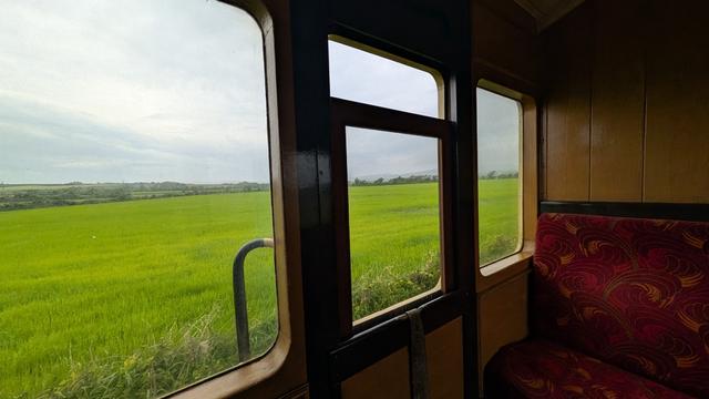 A view out of a wooden train carriage's windows, looking over green fields with cloudy sky. There is an empty seat on the opposite side of the compartment.