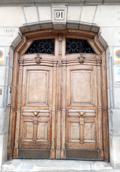 A weathered wooden set of double doors with a metal filigree panel above each  and a plain but element design.