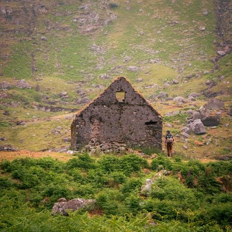A man walks towards and abandoned stone structure in a green, rocky valley.