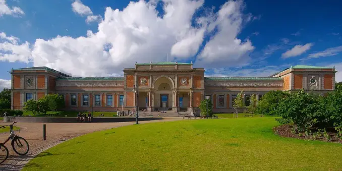 The image depicts a large building with impressive architecture, likely a museum or cultural institution. It features a broad façade with decorative elements, columns, and a central entrance, set against a blue sky with clouds. The surrounding area includes green lawns and trees, and there are a few people visible, along with a bicycle in the foreground.

Image Credits: Wikimedia / Jiří Komárek / CC BY-SA 4.0