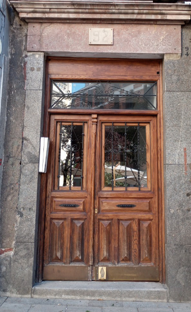 A well maintained normal sized doorway with a varnished wooden door with glass and decorative metal panels at the top and mid-top of the door